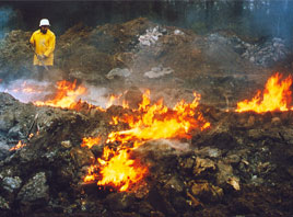 Offener Deponiebrand infolge chemischer Reaktionen. Es sind Flammen und Qualmm zusehen. Ein Mitarbeiter im Schutzanzug steht am linken Rand